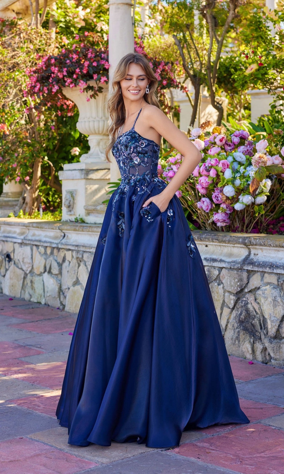 A woman wearing the Long Prom Dress: Juliet JT2617U in navy blue stands outdoors near blooming flowers and a stone railing.